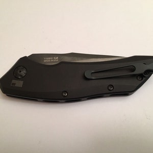 May include: A black folding knife with a pocket clip and a black handle. The knife has a silver blade and the text "7100BW CAI MADE IN USA" is engraved on the handle.