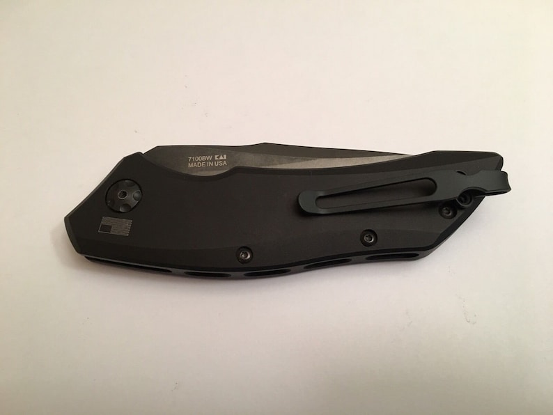 May include: A black folding knife with a pocket clip and a black handle. The knife has a silver blade and the text "7100BW CAI MADE IN USA" is engraved on the handle.