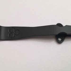 May include: Black metal pocket clip with a three-circle logo. The clip has two holes on the side.