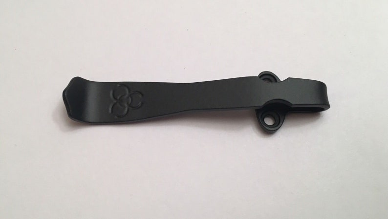 May include: Black metal pocket clip with a three-circle logo. The clip has two holes on the side.