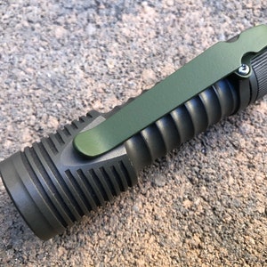 Puede incluir: Una linterna verde oscuro con un cuerpo texturizado y un clip de bolsillo verde. La linterna tiene un tornillo negro en el extremo.