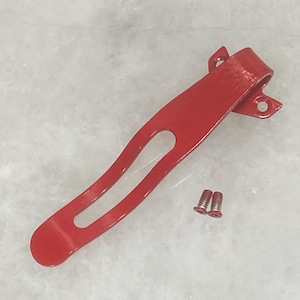 May include: A bright red metal clip with a curved design and two small screws. The clip has a rounded end and a hole in the center. The clip is likely for securing items.