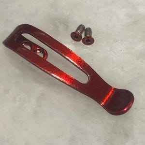 Könnte beinhalten: Ein roter Metall-Taschenclip mit zwei silbernen Schrauben. Der Clip ist so konzipiert, dass er an einer Tasche oder einem Gürtel befestigt werden kann.