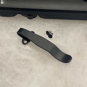 May include: Black metal pocket clip with two silver screws. The clip is designed to attach to a pocket or belt.