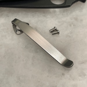 May include: A silver pocket clip for a knife. The clip has a rounded end and two holes for screws. Two small silver screws are included.