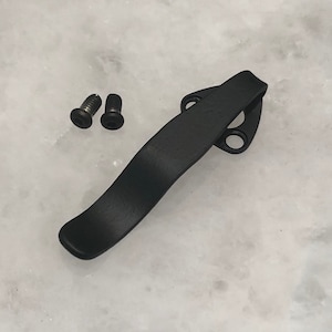 Könnte beinhalten: Ein schwarzer Metall-Taschenclip mit zwei kleinen Schrauben. Der Clip hat ein gebogenes Design mit zwei Löchern an einem Ende zur Befestigung. Die Schrauben sind schwarz mit einem Sechskantkopf.