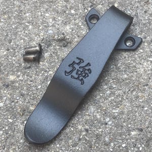 May include: A matte black metal pocket clip featuring a stamped Japanese kanji character on the surface. The clip has two circular mounting holes at the top and is accompanied by two small silver-colored metal screws positioned to the left side.