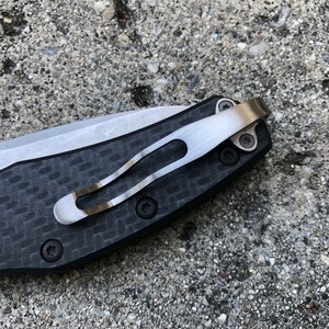 May include: Close-up of a black and silver pocket knife with a carbon fiber handle and a clip. The clip is attached to the handle with two screws.