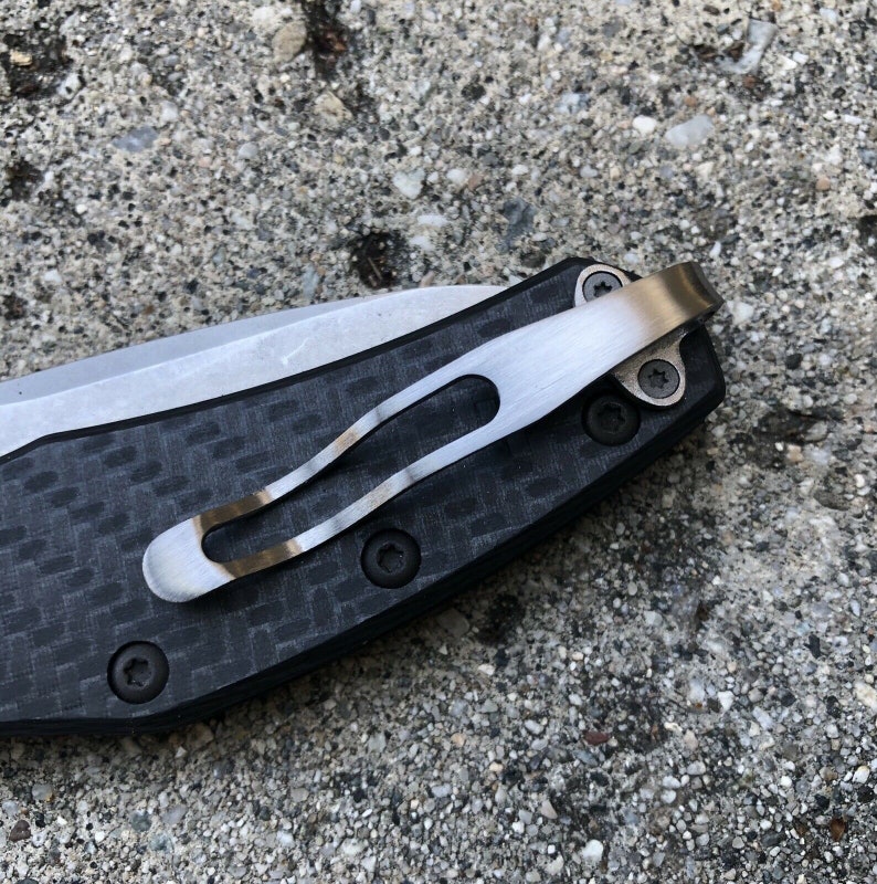 May include: Close-up of a black and silver pocket knife with a carbon fiber handle and a clip. The clip is attached to the handle with two screws.