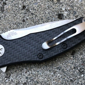 May include: A black and silver pocket knife with a carbon fiber handle and a clip. The knife has a small star-shaped screw on the handle and the text "0770CF KAI-manufactured MADE IN USA" is engraved on the blade.