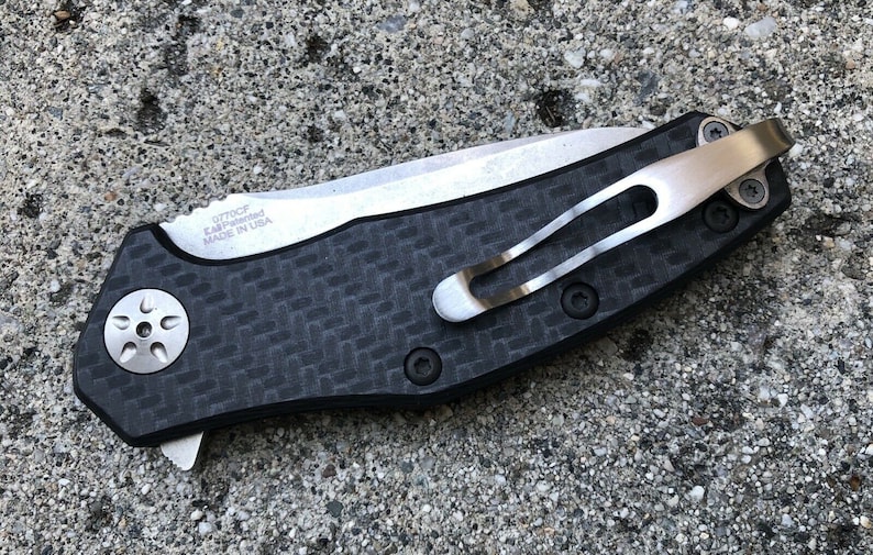 May include: A black and silver pocket knife with a carbon fiber handle and a clip. The knife has a small star-shaped screw on the handle and the text "0770CF KAI-manufactured MADE IN USA" is engraved on the blade.