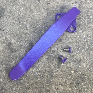 May include: A purple metal pocket clip with a triangular mounting base featuring two circular screw holes, accompanied by two small matching purple screws with flat heads, designed for attachment to a tool or accessory.