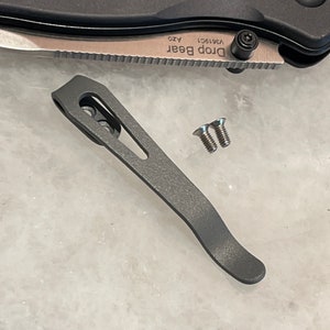 May include: A grey titanium pocket clip for a knife. The clip has a curved design and two small screws. The clip is designed to attach to a pocket or belt.