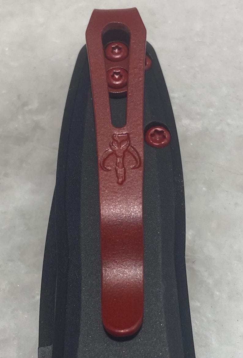 May include: A black pocket knife with a red clip and a Mandalorian symbol. The clip has three screws.