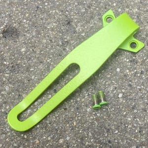 May include: A lime green metal pocket clip featuring a long, narrow cutout slot and two mounting holes at the top, accompanied by two small matching lime green screws with silver-colored threaded shafts for secure attachment to a tool or device.