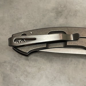 May include: A close-up of a silver pocket knife with a clip. The clip is attached to the knife with two screws.