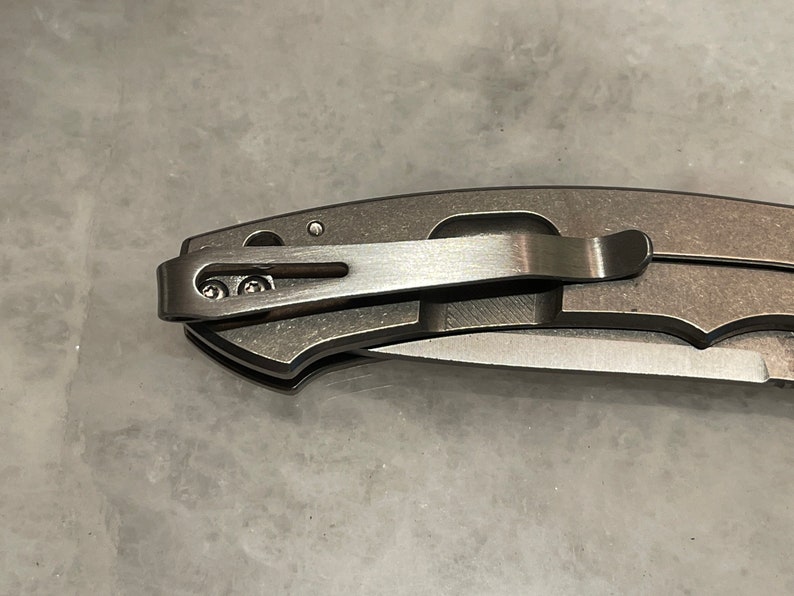 May include: A close-up of a silver pocket knife with a clip. The clip is attached to the knife with two screws.