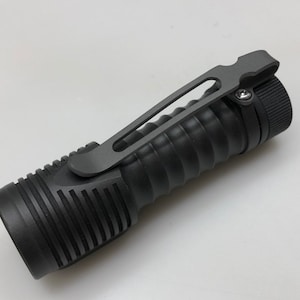 May include: A black flashlight with a textured grip and a pocket clip. The flashlight has a black metal body and a black plastic head.