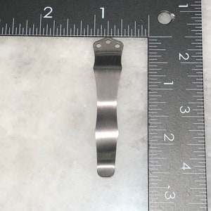 May include: A silver metal pocket clip with three holes at the top. The clip is about 2 inches long and is designed to attach to a pocket or belt.
