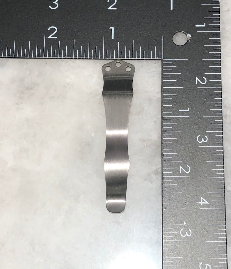 May include: A silver metal pocket clip with three holes at the top. The clip is about 2 inches long and is designed to attach to a pocket or belt.