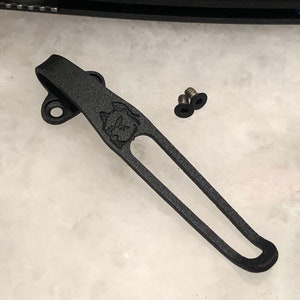 Könnte beinhalten: Schwarzer Metall-Taschenclip mit einem kleinen, gravierten Tiermotiv. Der Clip hat ein abgerundetes Ende und ein kleines Loch oben.