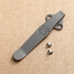 May include: A dark gray metal pocket clip featuring an embossed paw print design on the surface. The clip has a mounting base with two circular screw holes and is accompanied by two small, separate silver-colored metal screws for attachment.