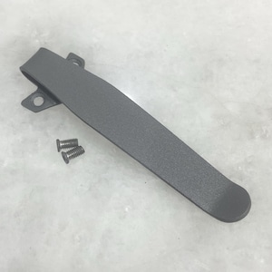 May include: A gray pocket clip with two small screws. The clip is made of metal and has a curved design with a rounded end. It features two holes for attachment and is designed to secure items.