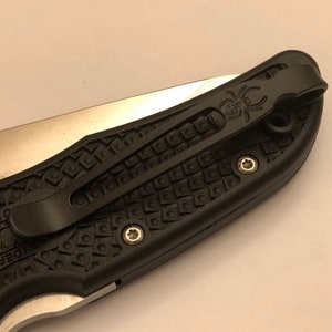 May include: Black pocket knife with a textured black handle and a silver blade. The knife has a black pocket clip with a spider logo.