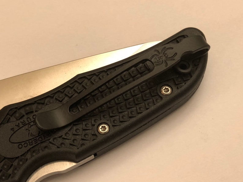 May include: Black pocket knife with a textured black handle and a silver blade. The knife has a black pocket clip with a spider logo.