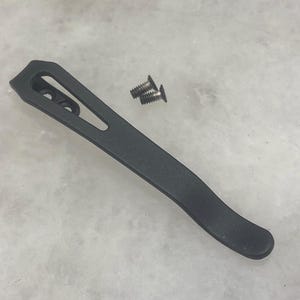 May include: A black metal pocket clip with two small screws. The clip has a curved design with a slotted opening and two screw holes. The clip is likely for attaching to a knife or other tool.