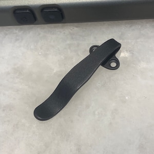 May include: A black metal pocket clip with a curved design and two holes. The clip is designed to attach to a pocket or belt.