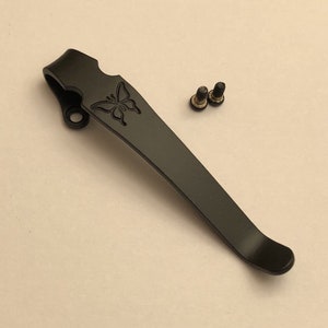 May include: Black metal pocket clip with a butterfly design. The clip is attached to a knife or multi-tool with two screws.
