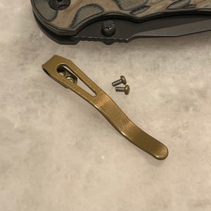 Może przedstawiać: Złoty zaczep do kieszeni do noża. Zaczep jest wykonany z metalu i ma zakrzywiony kształt. W zestawie znajdują się dwa małe śruby.