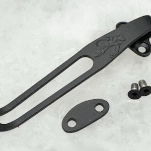 May include: Black metal pocket clip with a scorpion design. The clip is attached to a black metal oval with two holes. Two small black screws are included.