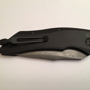 May include: A black pocket knife with a partially serrated blade and a pocket clip. The knife has a black handle with a textured surface and a small, round button on the side. The blade is partially open and the knife is lying on a white surface.