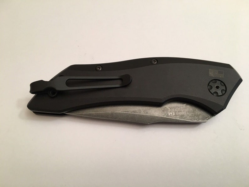 May include: A black pocket knife with a partially serrated blade and a pocket clip. The knife has a black handle with a textured surface and a small, round button on the side. The blade is partially open and the knife is lying on a white surface.