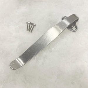 May include: A brushed metal pocket clip with two small screws. The clip is curved and has two holes for mounting. The clip is approximately 4 inches long.