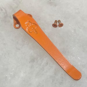 May include: A bright orange metal pocket clip featuring an embossed skull-like emblem near the top mounting point, accompanied by two small matching orange screws with star-shaped drive heads for secure attachment to a folding knife handle.