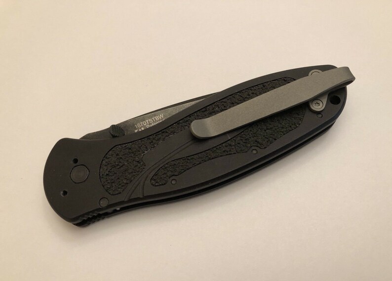 May include: Black pocket knife with a textured black handle and a silver pocket clip. The knife has the text "1070TSBW" engraved on the handle.