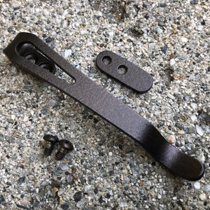 May include: A dark brown metal pocket knife with a curved blade and a small metal plate with two holes. The knife is on a gray and brown gravel surface.