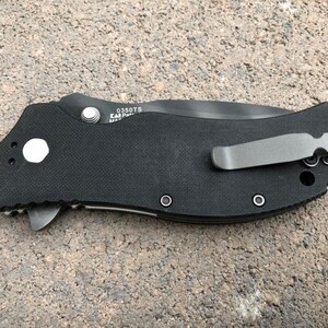 May include: Black pocket knife with a black handle and a silver pocket clip. The knife has a textured handle and a silver button on the side. The knife has the text "0350TS" and "EAR" on the side.