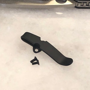 May include: Black metal pocket clip with two small black screws. The clip is designed to attach to a pocket knife.