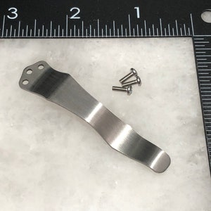 May include: A silver pocket clip with three holes at the top and a curved shape. The clip is shown with four small silver screws.