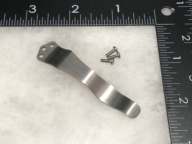 May include: A silver pocket clip with three holes at the top and a curved shape. The clip is shown with four small silver screws.