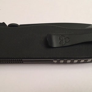 May include: Black pocket knife with a black clip and a textured black handle. The knife has a silver blade and a black and silver patterned design on the back.