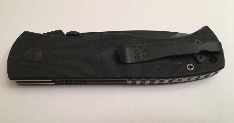 May include: Black pocket knife with a black clip and a textured black handle. The knife has a silver blade and a black and silver patterned design on the back.