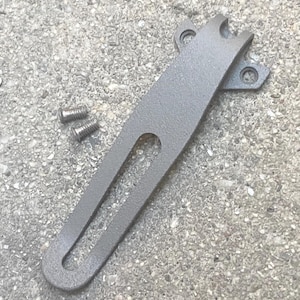 May include: A matte gray metal bracket featuring an elongated central slot and a top section with two circular mounting holes. The bracket has a textured finish and is accompanied by two small, threaded metal screws positioned to the left side.
