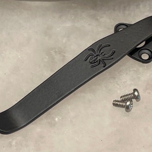 May include: Black metal pocket clip with a spider design and two silver screws.