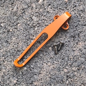 Puede incluir: Un clip de bolsillo de metal naranja con dos tornillos. El clip está diseñado para sujetarse a un bolsillo o cinturón.