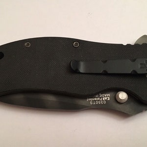 Pu&ograve; includere: Coltello pieghevole nero con un fermaglio da tasca nero e un logo a forma di teschio. Il coltello ha un'impugnatura nera e una lama argentata. La lama &egrave; parzialmente aperta e il coltello &egrave; chiuso. Il coltello ha il testo "03501S KAI Patented MADE IN" sul lato.